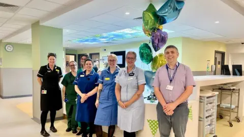 Medical professionals - all but one in uniform - lined up in front of a hospital reception area with balloons behind them. All are smiling at the camera.