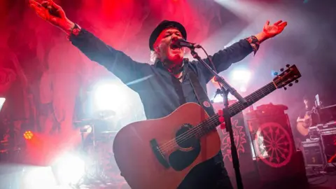 A man singing into a mircrophone on stage, with a guitar. 