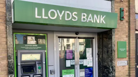 A branch with the words Lloyds Bank in white above the entrance. A cash machine is on the left and there are doors in the middle.