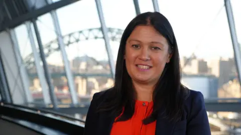 Lisa Nandy standing inside the Glasshouse in Gateshead. She has dark long hair and is wearing a red top and dark blazer. The Tyne Bridge can be seen through the windows behind her.