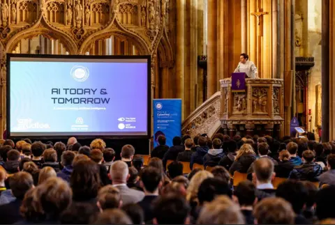 TechSPARK A man stands in the lecturn at Bristol Cathedral in front of dozens of young people, with a very large screen at the front of the nave with a slide saying 'AI Today & Tomorrow'