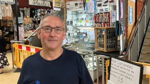 Photograph of Jonathan Lamb, who runs a watch repair business at Wigan indoor market. The image shows him in front of his store. He has grey hair and wears glasses and a navy blue t-shirt.