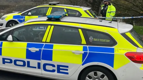 Two stationary police cars on a road. A police officer is standing behind one next to police tape.