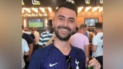 Essex Police A man smiles at the camera. He is wearing a football shirt which is navy blue and is holding a bottle. He has short dark hair and a dark short beard.