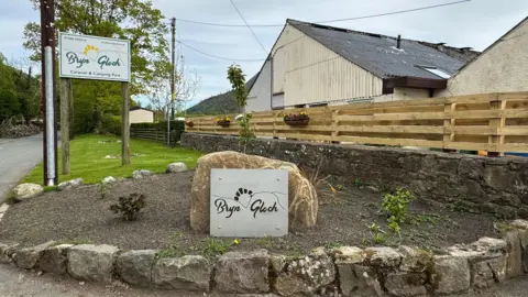 Entrance to Bryn Gloch campsite with sign against a boulder with name of campsite and another sign behind and higher up on the side of the road. Buildings are visible to the right and the road to the left