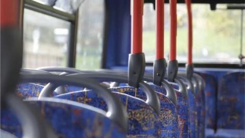 An image showing blue bus seat on the top deck of a double-decker bus. There are orange metal bars coming from the top of each seat, attaching to the ceiling. It is daytime outside the windows. 