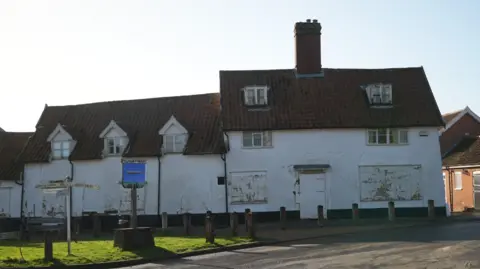 The whitewashed former pub, which is made up of two adjoined buildings. One is three storeys and the second is two storeys. Its ground floor windows are boarded up. The village sign stands on a green space outside the pub.