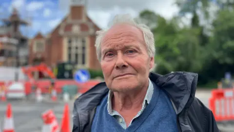 Graham Browne standing in front of the roadworks which are orange cones and fencing. behind him is the old courthouse which lights will be placed in front of. he has a rain coat on and navy blue jumper and gray hair