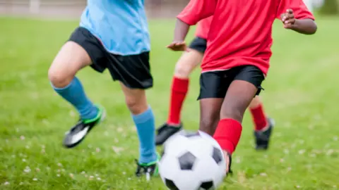 A stock image showing three children running on grass and a football in the foreground. Two of the children are in red tops and black shorts while the third is in a blue top and black shorts.