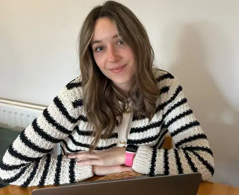Abbi Lily A woman in a black and white striped cardigan sits at a table indoors, with her arms folded. She smiles at the camera.