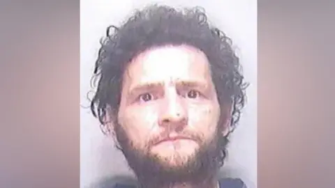 A police mugshot of a man with untidy black hair and a short beard. He is staring directly into the camera.