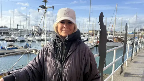 Lili Sheppard Julie Forsyth in a cap and jacket, smiling for the camera. Several yachts in Ramsgate Harbour are seen in the background.