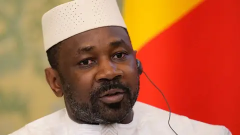 Mali's junta leader attends a meeting with Russia's President at the Kremlin in Moscow. He wears a white outfit and communicated with the help of a translation earphone. Behind him is the national flag of Mali.