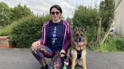 A woman in sunglasses, a purple sweater, dark tee-shirt and shiny scale-pattern leggings kneels next to a German Shepherd dog on a path in front of a hedge. 
