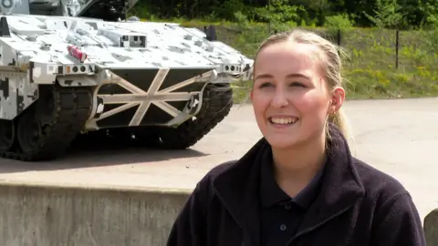 A blonde haired women wearing a black fleece stands in front of an armoured vehicle