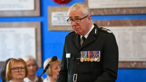 PSNI chief constable Jon Boutcher, standing in front of a small microphone. He is wearing a black suit with medals on the front and glasses. There are women sitting to the left of him. 