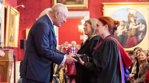 Two women from Hull College receive the award from King Charles III in a red panelled room with an audience sitting behind them
