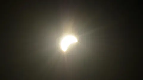 BBC Weather Watchers / Tony_L A close-up photograph shows the Sun during the eclipse as a bright crescent shape against a dark sky in Worthing, West Sussex. Rays of light extend from the Sun, while part of its disc is obscured by the Moon.