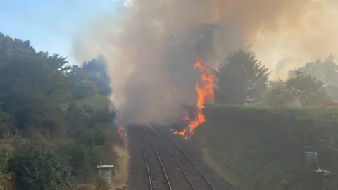 Flames tower above a large hedgerow on the right side of a railway line. A huge plume of smoke completely blocks the view up the line.