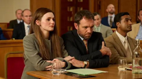 Rob Baker Ashton / Channel 4 A picture of a man and woman sat at a desk. They are sitting in a room with a number of other people around them. 