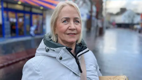 A woman with light hair looking at the camera, she is wearing a grey coat which is zipped up to the neck. She is standing on a high street with shops in the background. 