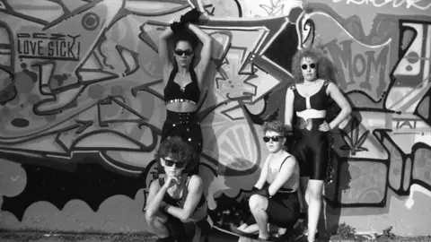 Sharon Hepburn Women stand in front of a graffiti‑covered wall, wearing dark outfits and sunglasses.
