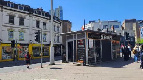 Bristol City Council This picture was taken in Bristol City Centre near the Bristol Shopping Quarter sails structure. It has a picture of a black food kiosk. It is the Cornish Bakehouse kiosk. The shutters for the kiosk is closed. People are standing around the shop waiting to cross the road. 