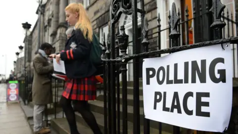 A young woman wearing a red and black tartan skirt leaves a polling station in Edinburgh, Scotland, during the 2014 independence referendum. A sign reading: Polling place is attached to black railings.