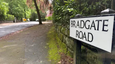 BBC Street view of a road sign reading: 'Bradgate Road'. The sign is small, and white with black writing. It is positioned on a pavement 