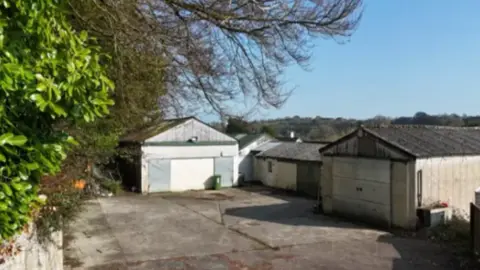 Torridge District Council A small yard surrounded by several old, single‑storey industrial outbuildings. The ground is a wide expanse of worn concrete with visible cracks and patches of discolouration. On the right, there is a rectangular building with a corrugated roof and a large garage-style door. Further back, there are two more outbuildings with pale walls and sloping roofs, one of which has a green wheelie bin positioned outside.