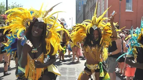 Two women wearing bright yellow feathered headdresses and bejewelled bikini tops, dancing in the street during St Paul's Carnival. It is a sunny day and they are surrounded by people.