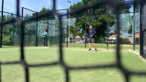 Two people on a court playing padel. They are holding racquets and there is a net in the foreground of the shot which is blurred in the picture