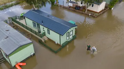 Getty Images People wade through floodwater surrounding mobile homes at Cogenhoe Mill Holiday Park after the River Nene burst its banks, on September 24, 2024 in Cogenhoe, England