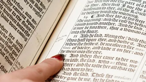 Duggleby Stephenson A woman's finger with a painted red finger nail points onto a page of a large old book with copper plate printed words on old yellowed paper.
