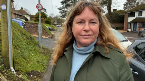 BBC Woman with brown hair and blue turtleneck top stands on a pavement on a residential road. There are cars parked to the right. There is a circular sign which says '20mph' attached to a lamp post.
