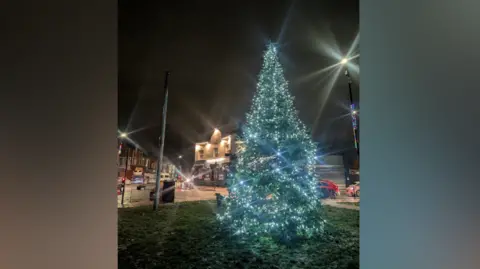 Friends of Fulwell The Fulwell Christmas tree is tall and has a string of blue and yellow lights. It is placed on a grassed area near a road. There are some houses across the street.