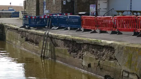 LDRS A crumbling concrete wall and footpath alongside the harbour. There are large chunks of concrete missing from the wall. Thee path is cordoned off with plastic fencing panels. The tide is in and the water is about a metre below the path.