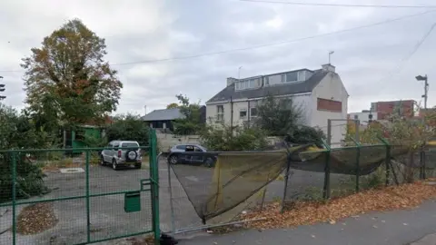 The image shows a fenced off car park with two cars parked inside. Houses border the pitch to the right.