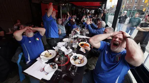 Getty Images A bar table full on Italy fans, in blue team jerseys, react as their team scores a goal