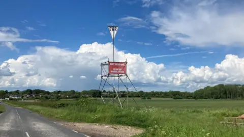 Mark Williamson/BBC Sign on a pylon saying East West Rail is on the wrong track in a field beside the road