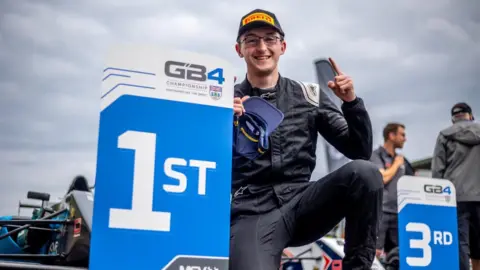 Mat Acton Lucas Blakeley has his finger pointed in the air. He has a 1st place sign next to him as he celebrates winning a race.