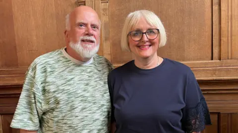 Leslie Judd  and Sharon Fox stand next to each other in front of wood panelling. 
Leslie wears a green and white t-shirt and has white hair and a beard and a nose ring. Sharon is wearing a navy blue top and has a blond bob and black glasses.