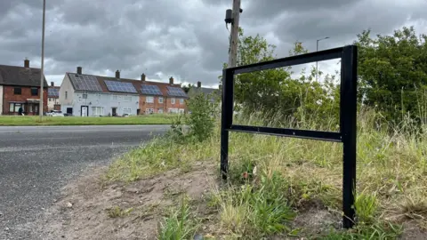 A black metal frame where the Dungeon Lane road sign would have been on a grass mound. A main road runs alongside the lane with an terraced houses and a telegraph pole in the background.