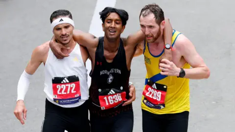 Three men wearing black shorts and different coloured running vests jog towards the finish line of the Boston Marathon with their arms around each other. Robson De Oliveira, who has short, dark hair and a moustasch and is wearing a white vest and a white headband. Ajay Haridasse has short, dark hair and a black vest. Aaron Beggs has short brown hair and a beard and is wearing a yellow vest with a blue stripe. 