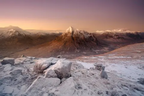 Cliff Hands A wide view of a snow‑dusted mountain range at sunrise, with soft pink and golden light illuminating a steep central peak and frosty rocks in the foreground.