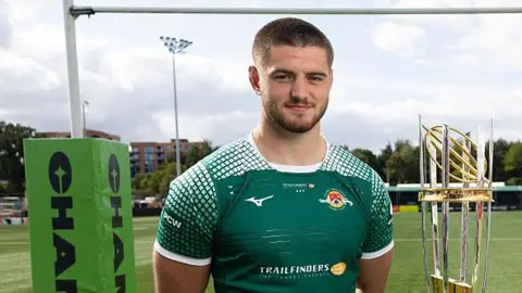 Ealing Trailfinders captain Josh Taylor on the left poses in front on the Champ winners trophy with rugby posts in the background