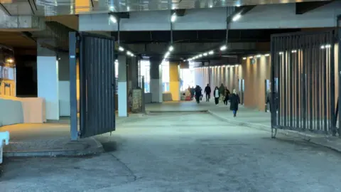 A wide covered passageway stretches ahead, lit by rows of ceiling lights. Several people walk through the space, heading in different directions. Large metal gates stand open on either side of the entrance.