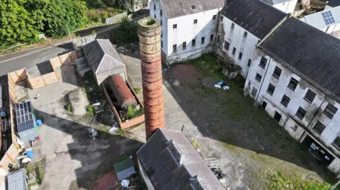 An old mill site with a large brick chimney and other crumbling buildings around it