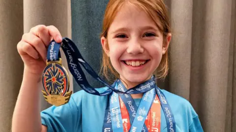 Marie Wood A young girl wearing a blue t-shirt, smiling proudly at the camera, holding a British Transplant Games medal with five other medal ribbons around her neck.