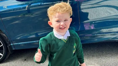 Kent Police Peter Maughan, a four-year-old boy with short, blond hair, is standing by a blue car wearing a school uniform. He has a white shirt, a green and yellow tie and a green jumper. He is holding a green schoolbag and is making a thumbs-up sign.